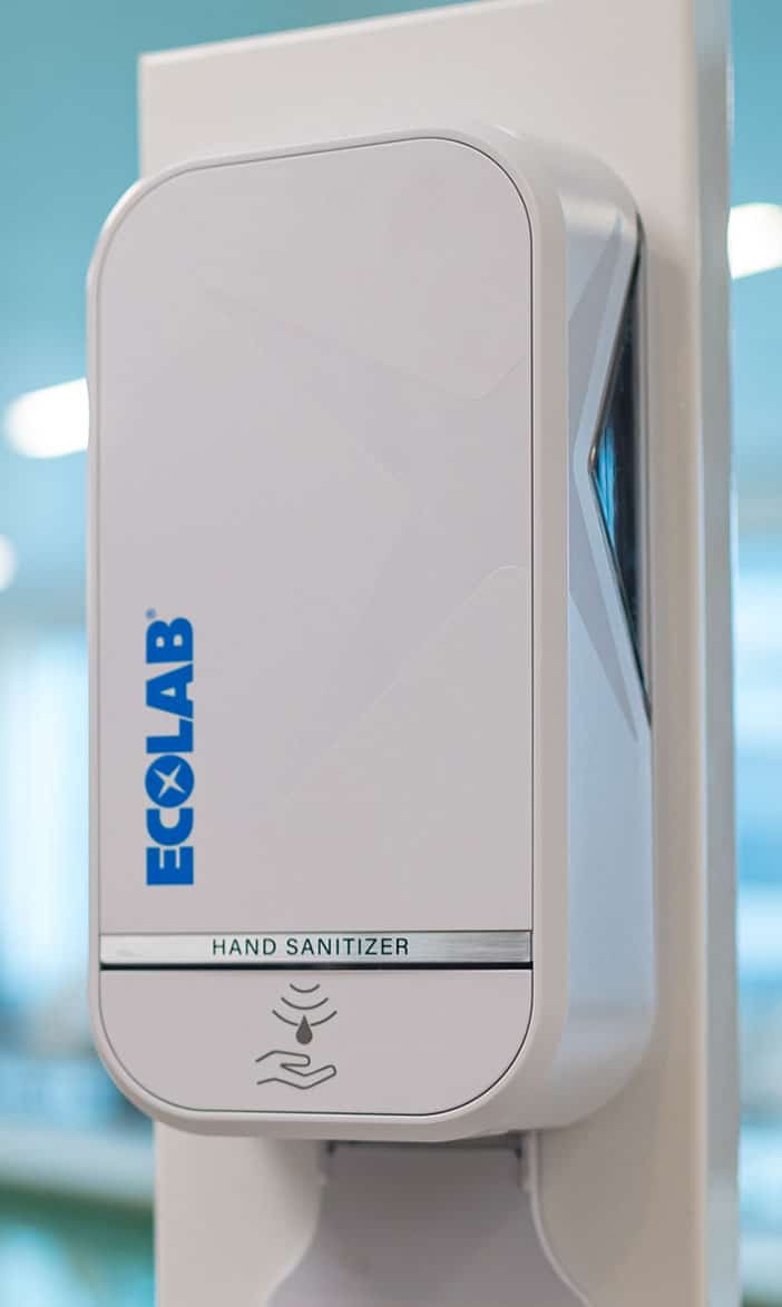 Side view of a white Ecolab touch-free hand sanitizer dispenser mounted on a wall in a blurred office or healthcare hallway.